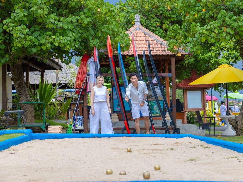 Two people playing bocce in a sand play area surrounded by greenery and a hut.