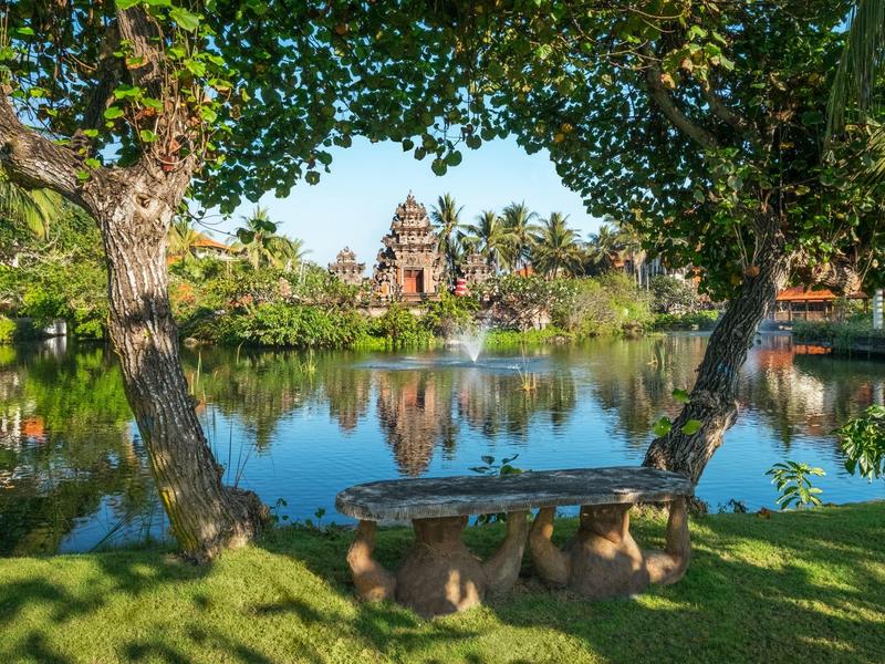 View of a pond with green lawn and ancient temple in the background under a blue sky.