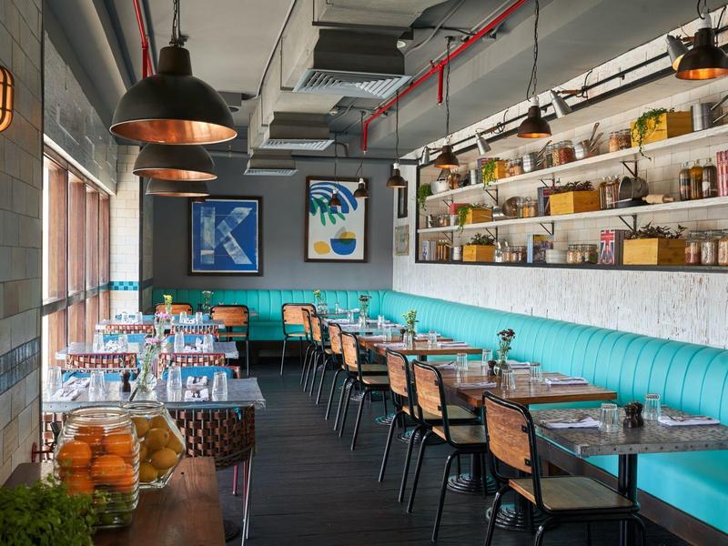 Ristorante moderno e luminoso con panca imbottita blu, sedie in legno e scaffali con bottiglie sul muro.