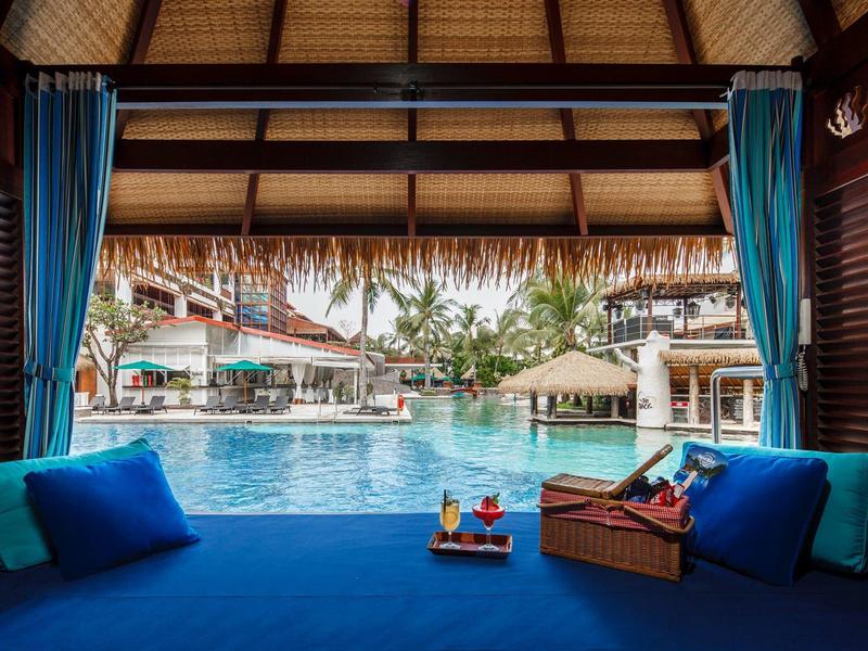 Vista da un lounge coperto a bordo piscina in un resort con cuscini blu e bevande.