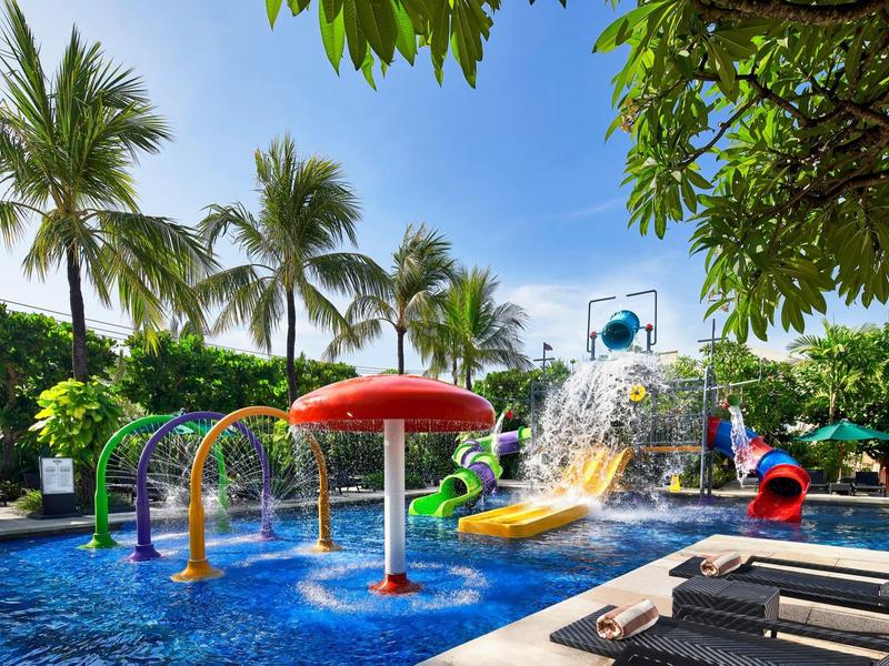 Area giochi acquatica colorata con scivoli e fontane nella piscina, circondata da palme e lettini.