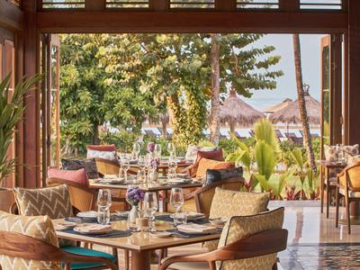 Offenes Restaurant mit Holzmöbeln und Blick auf tropische Pflanzen und Strohdach-Häuser.