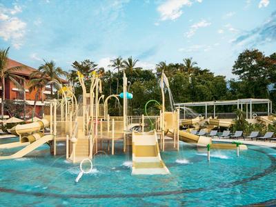 Hotelpool mit Wasserspielplatz, Rutschen und Liegestühlen unter blauem Himmel.