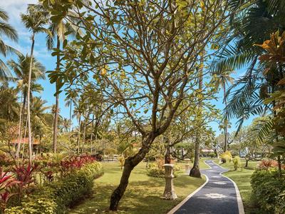 Ein gepflasterter Weg führt durch einen grünen tropischen Garten mit Palmen und blühenden Pflanzen.