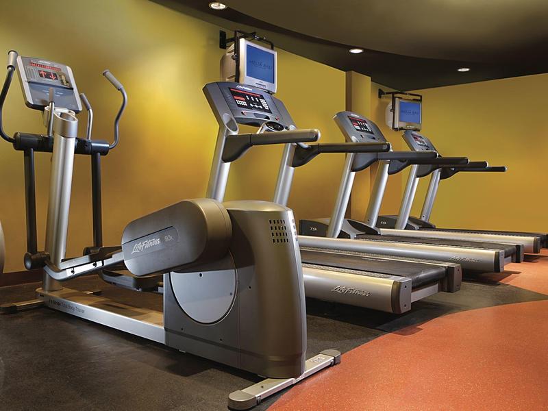 Fitnessraum mit drei Laufbändern und einem Crosstrainer auf braunem Boden vor gelber Wand.