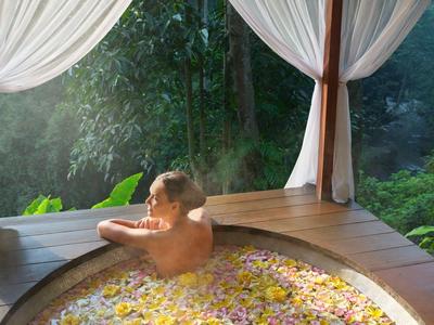 Eine Person entspannt in einem runden Holzbad voller Blütenblätter mit weißem Vorhang.