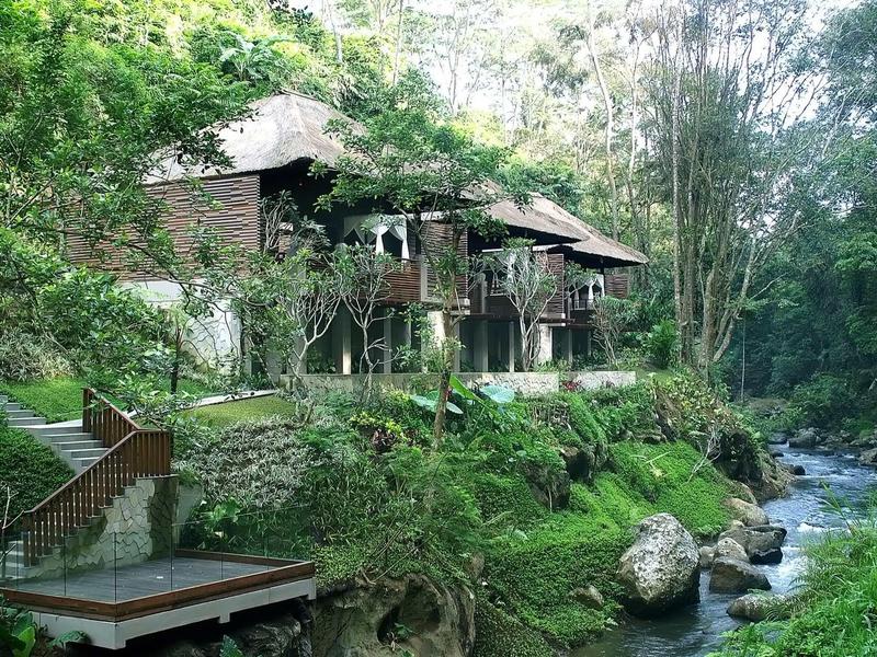 Altes Holzhaus am Fluss, umgeben von grünen Bäumen und Felsen am Ufer.
