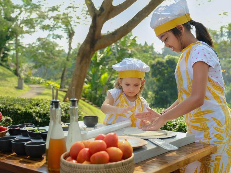 Zwei Kinder in Kochmützen bereiten draußen Teig vor, Tomaten und Flaschen stehen auf dem Tisch.