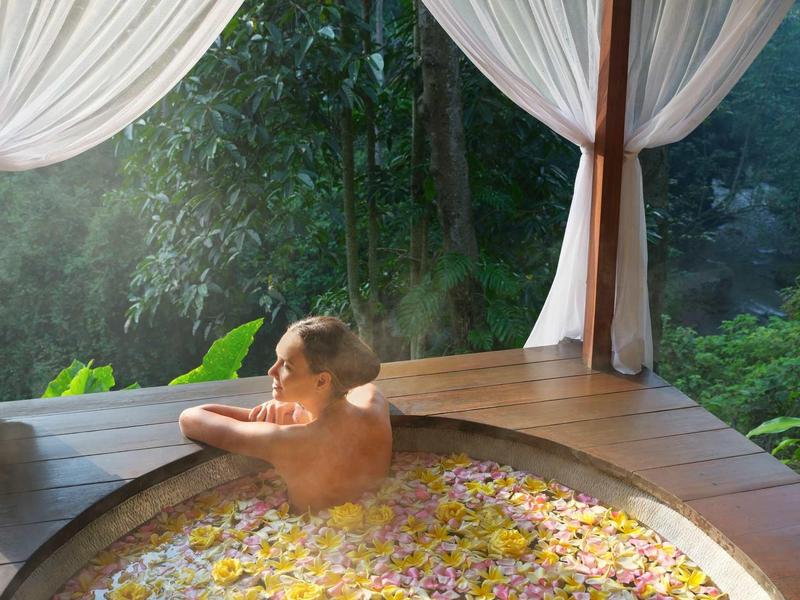 Eine Person entspannt in einem runden Holzbad voller Blütenblätter mit weißem Vorhang.