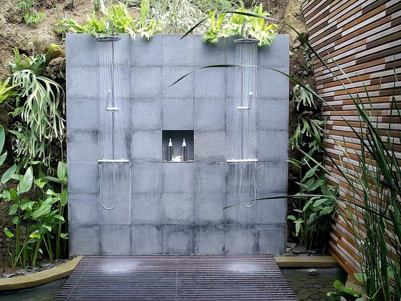 Moderner Wasserbrunnen mit quadratischer grauer Steinwand, umgeben von grünen Pflanzen und Holzpaneelen.