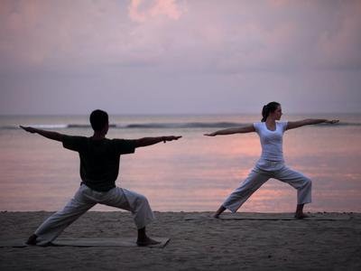 Zwei Personen praktizieren Yoga in Kriegerpose am Strand bei Sonnenuntergang.
