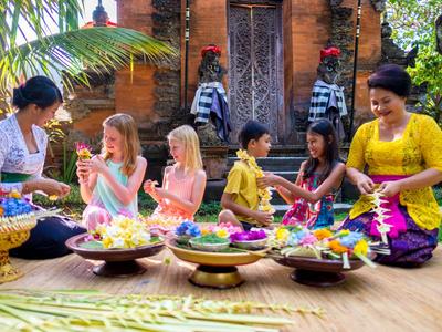 Frauen und Kinder bereiten in traditioneller Kleidung bunte Blütenarrangements vor, sitzend auf einem Boden vor einem Tempel.