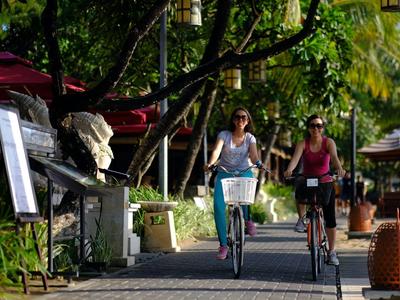 Zwei Frauen fahren auf einem schattigen Weg mit Bäumen und Polizeiwagen im Hintergrund Fahrrad.