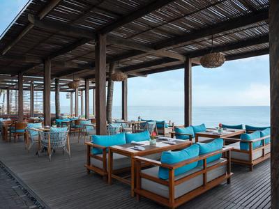 Open restaurantgedeelte met houten meubels en blauwe kussens met uitzicht op zee.