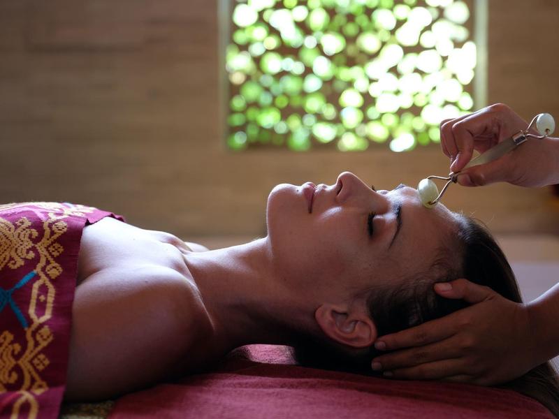 Vrouw krijgt ontspannende hoofdmassage in rustige spa met decoratief raam op de achtergrond.