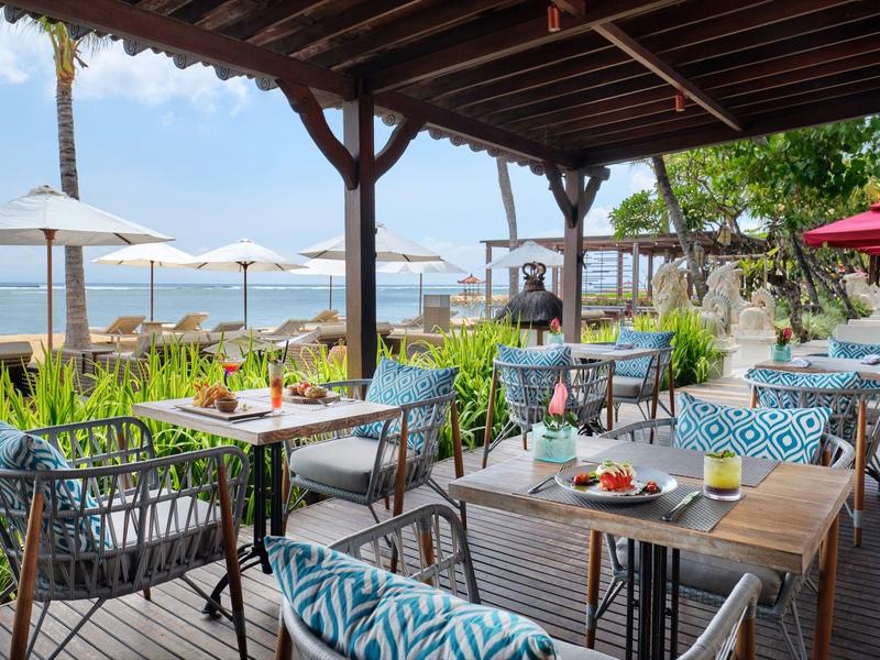 Overdekt buitenterras met comfortabele stoelen, tafels en uitzicht op zee.