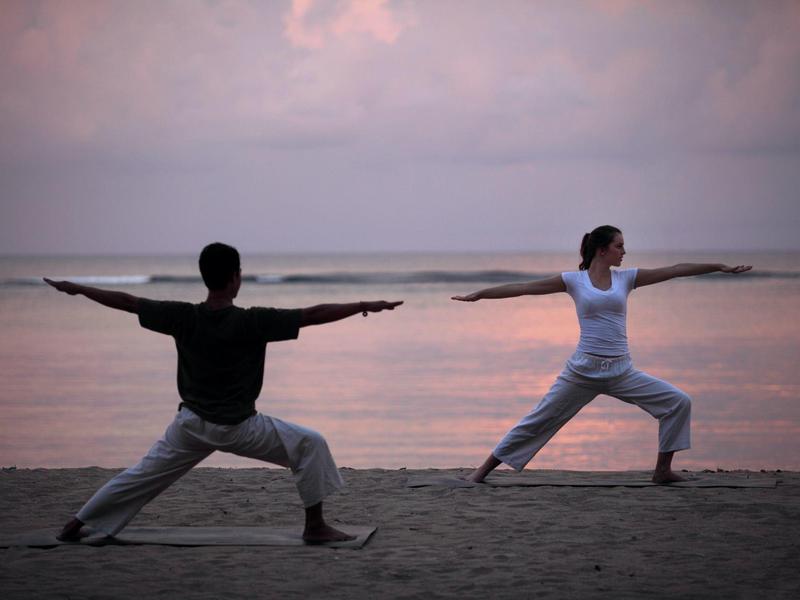 Twee mensen die de Warrior II-yogahouding doen op een strand bij zonsondergang.
