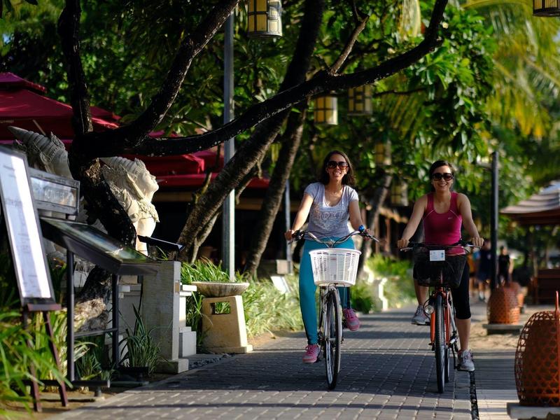 Twee vrouwen fietsen op een met bomen omzoomd pad omgeven door cafés.