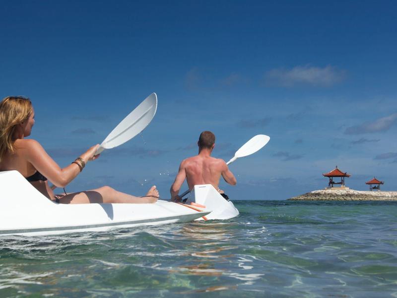 Twee personen peddelen in witte kajaks op helder water onder een blauwe lucht.
