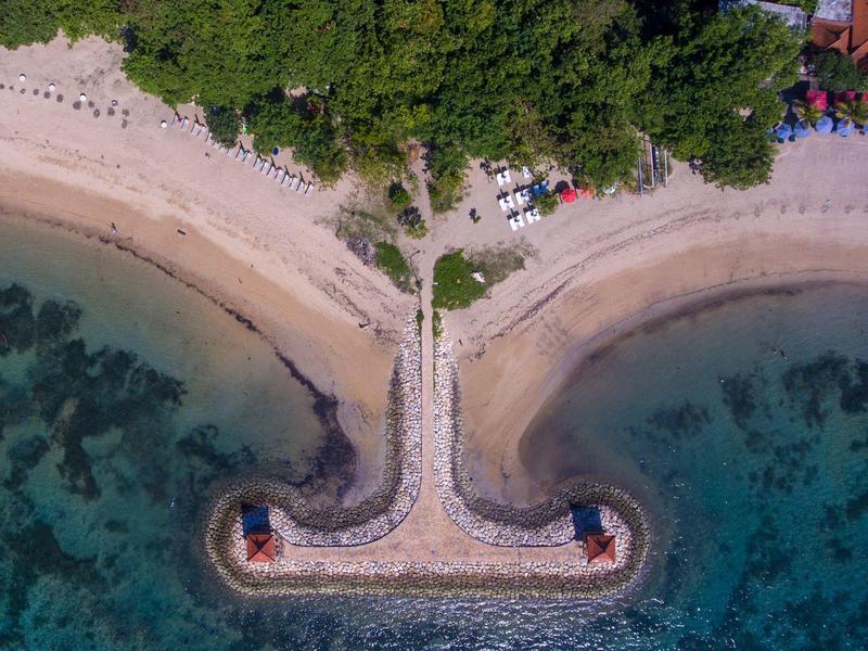 Luchtfoto van een betonnen pier met kleine torens die uitsteken in helderblauw water, omringd door zandstrand en groen gebladerte.