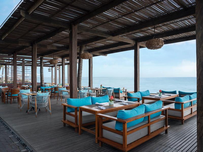 Open restaurantgedeelte met houten meubels en blauwe kussens met uitzicht op zee.