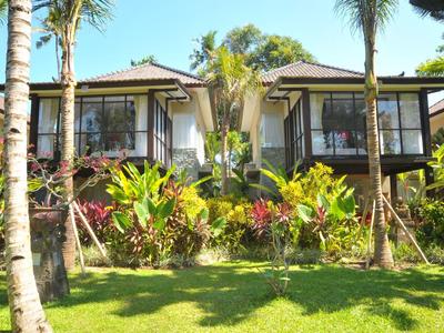 Dos villas de hotel con grandes ventanales, rodeadas de palmeras verdes y plantas sobre un césped soleado.
