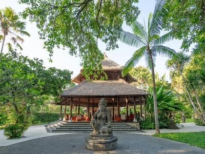 Un pabellón con techo de paja rodeado de árboles y una estatua de Buda de piedra en primer plano.