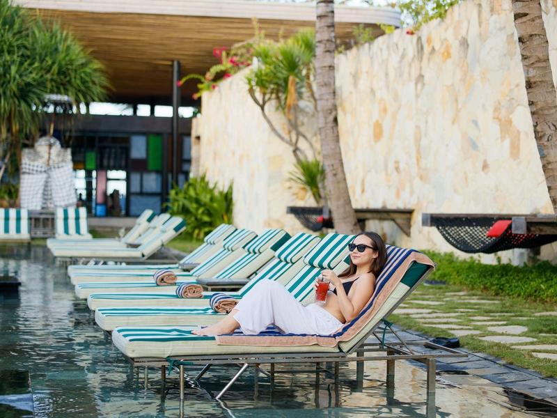 Mujer relajándose en una tumbona junto a la piscina del hotel con palmeras y hamaca al fondo.