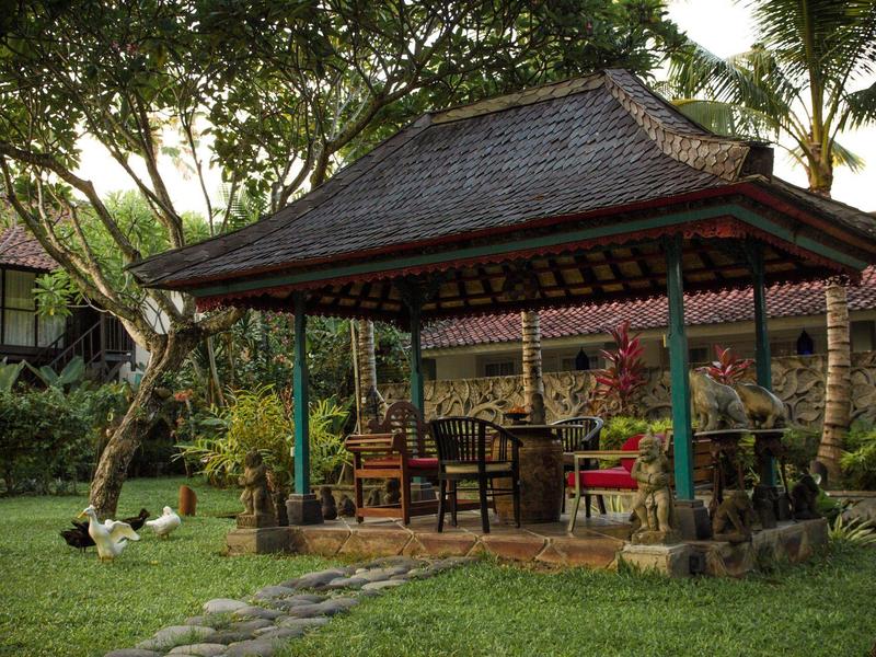 Cabaña de madera abierta con techo en un jardín verde con asientos y plantas.