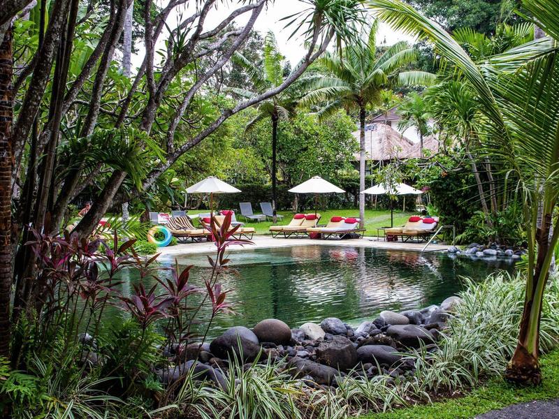 Estanque verde del hotel rodeado de palmeras y tumbonas con sombrillas.