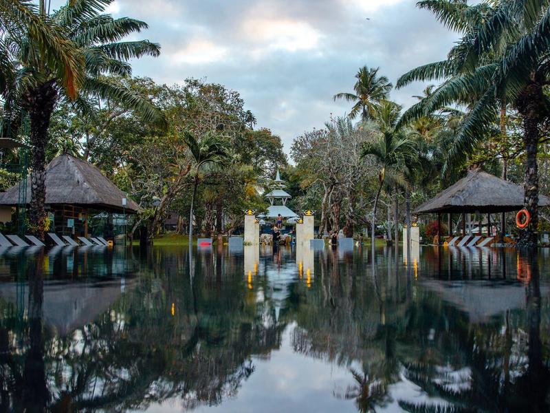 Vista de una piscina tranquila con palmeras y pequeñas cabañas bajo un cielo nublado.