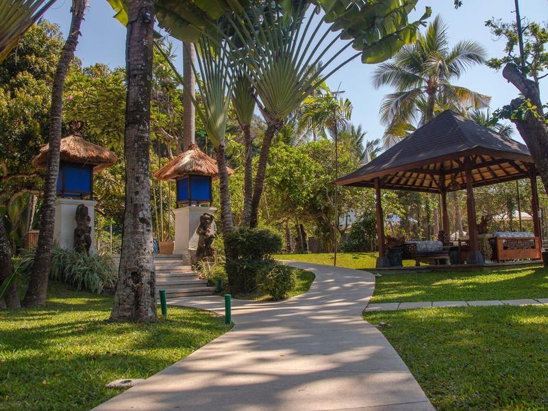 Camino pavimentado conduce a un pabellón y bungalows rodeados de palmeras en un jardín tropical de hotel.