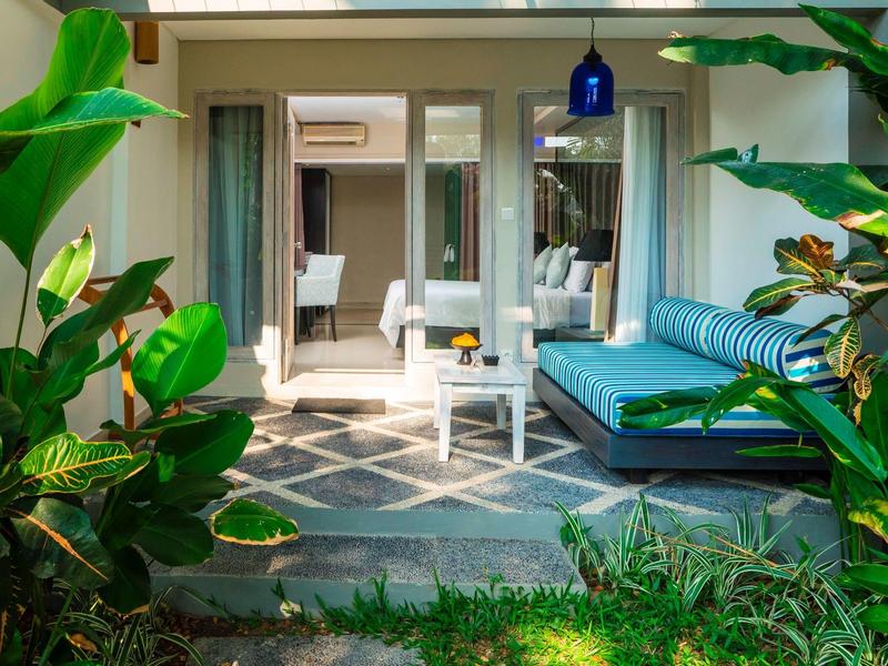 Vista de una terraza con sofá y mesa, rodeada de plantas verdes, frente a una habitación de hotel.