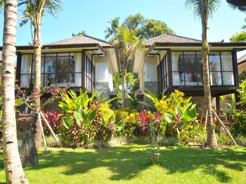 Dos villas de hotel con grandes ventanales, rodeadas de palmeras verdes y plantas sobre un césped soleado.