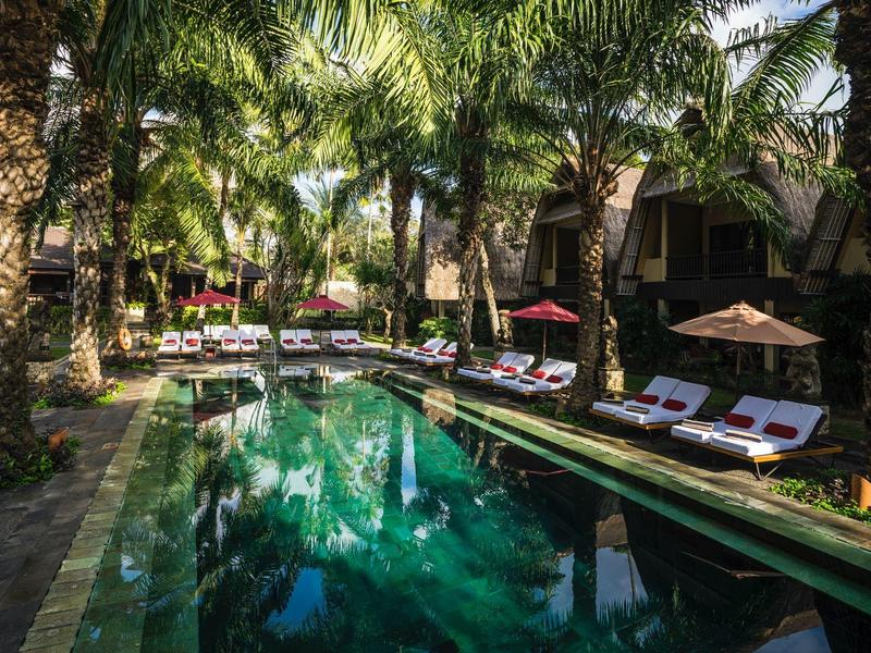 Piscina del hotel rodeada de palmeras con tumbonas y sombrillas junto a villas tropicales.