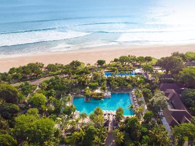 Luchtfoto van een resortzwembad omgeven door bomen op het strand met de zee op de achtergrond.