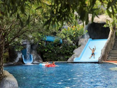 Kinderen glijden van een blauwe glijbaan in het zwembad, omgeven door bomen.