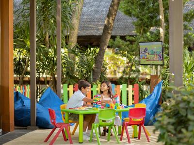 Kinderen zitten aan een kleurrijke buitentafel omringd door felgekleurde stoelen en groene vegetatie.