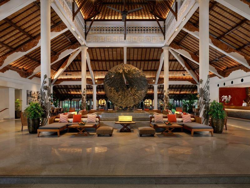 Ruime hotel lounge met hoog plafond, banken, planten en een decoratief kunstwerk in het midden.