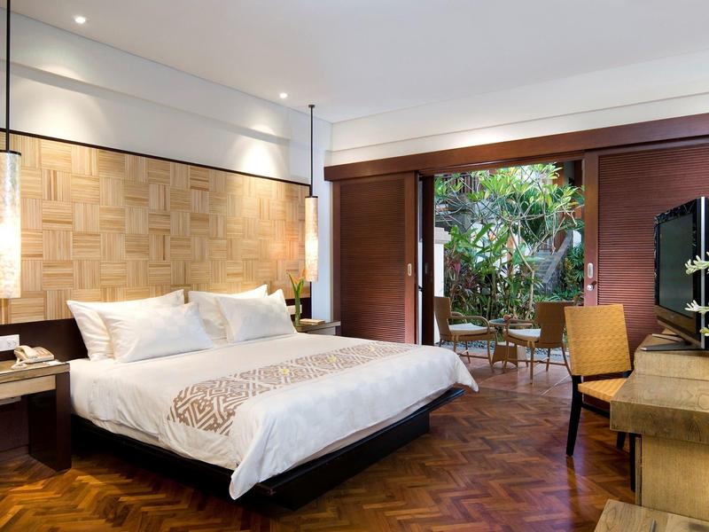 Lichte hotelkamer met een groot bed, houten vloer en toegang tot een terras met planten.