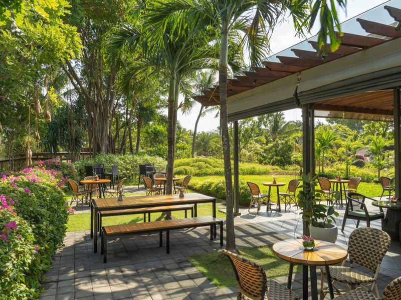 Buitenzitgedeelte met tafels en stoelen omgeven door weelderig groen en palmbomen.