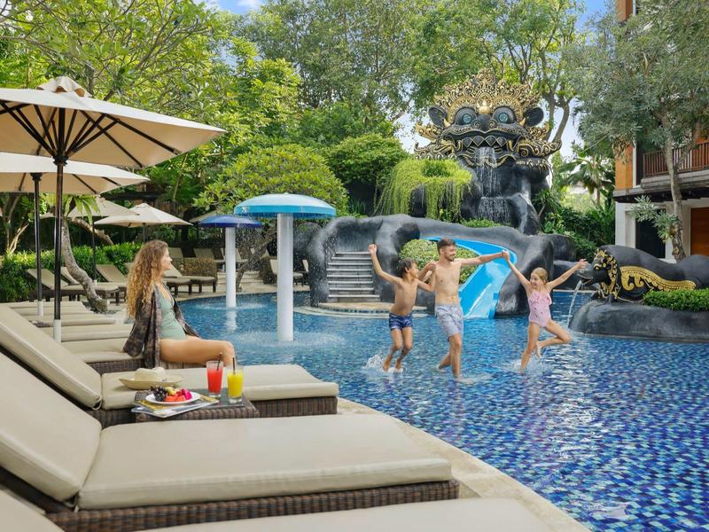 Kinderen spelen in een hotelzwembad terwijl een vrouw op een ligstoel onder parasols zit.