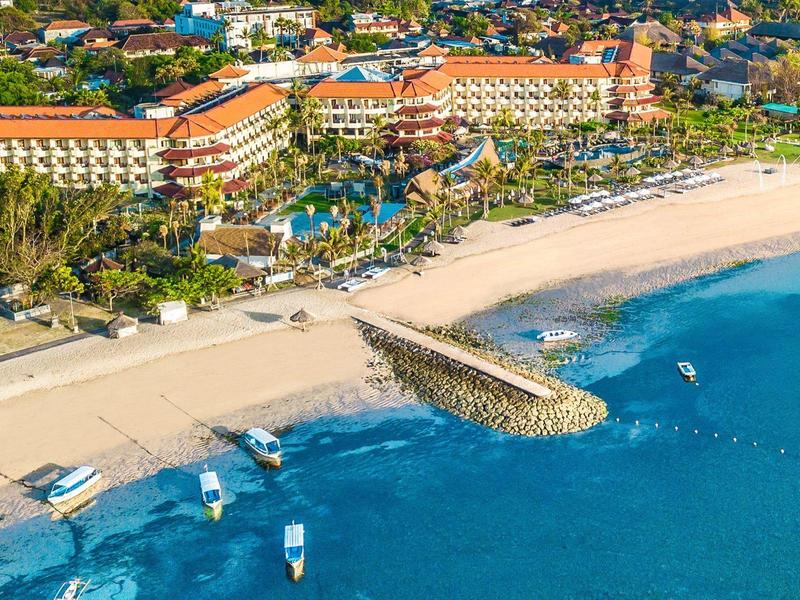 Hôtel en bord de plage avec piscine, chaises longues et bateaux dans une eau bleue claire.