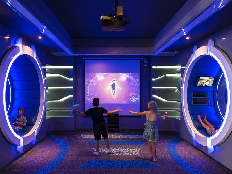 Des enfants jouent dans une salle futuriste avec des stations de jeu circulaires lumineuses et un grand écran.