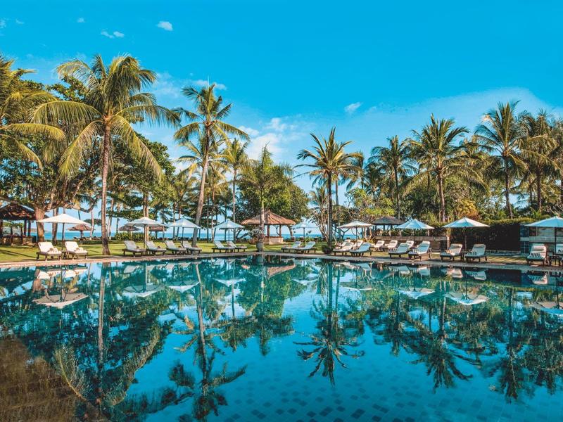 A serene pool surrounded by palm trees and lounge chairs under a blue sky.