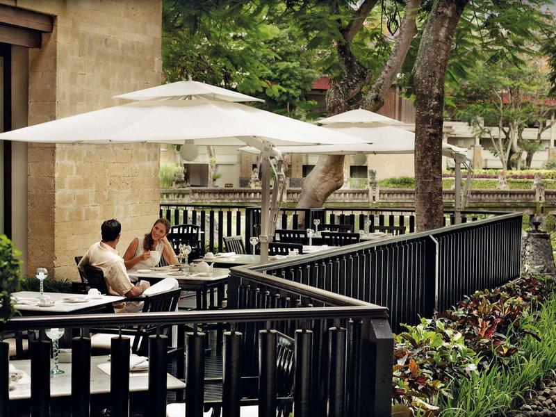 A man sits on a terrace with white umbrellas near a pond in a garden.