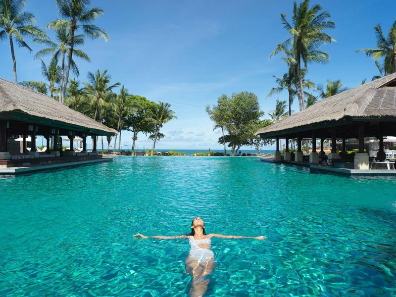 Woman relaxes in large pool between two covered lounging areas with palm trees.