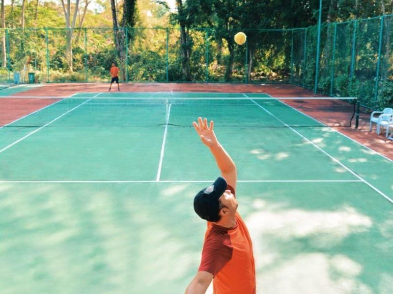 Man in red shirt serving a tennis ball on a green tennis court.