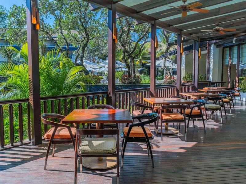 Covered veranda with several wooden tables and chairs in a quiet, green setting.
