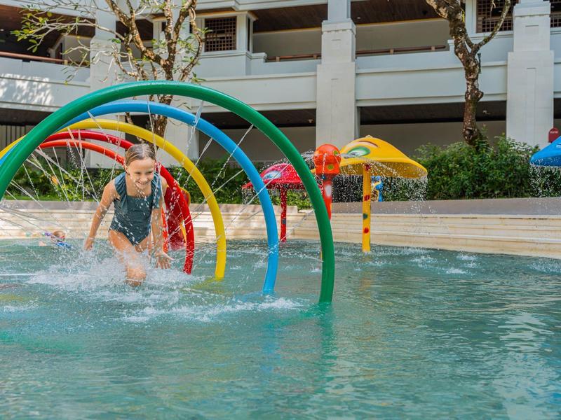 Child playing in a hotel outdoor pool with colorful water arches splashing water.